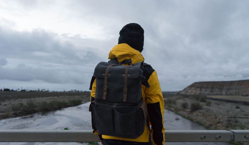 Figure in yellow jacket and black backpack stands facing a cloudy winter landscape.