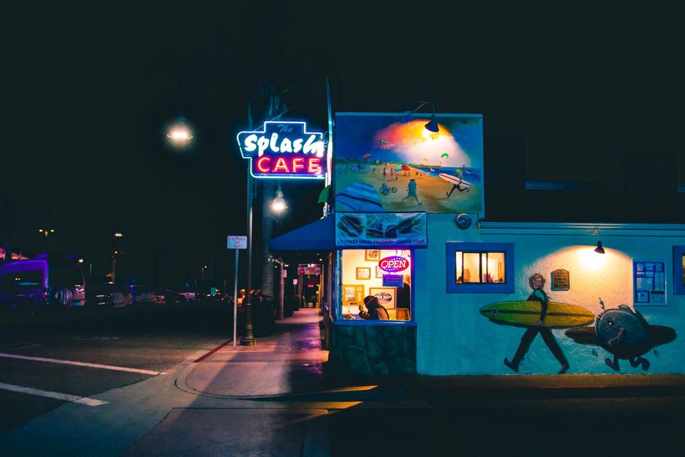 Retro neon signs illuminate a night scene outside a blue-lit diner.