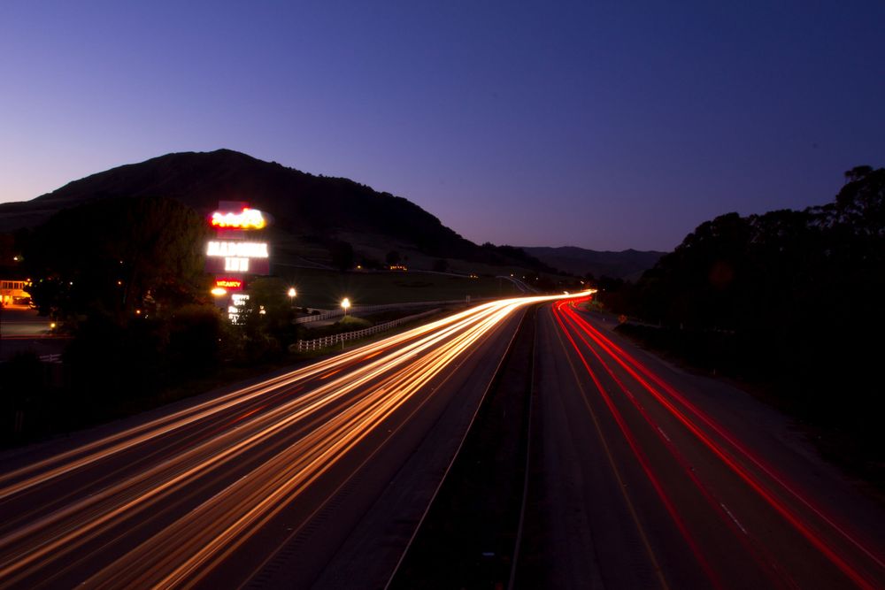 Long exposure of car light trails on highway at dusk with mountain silhouette.