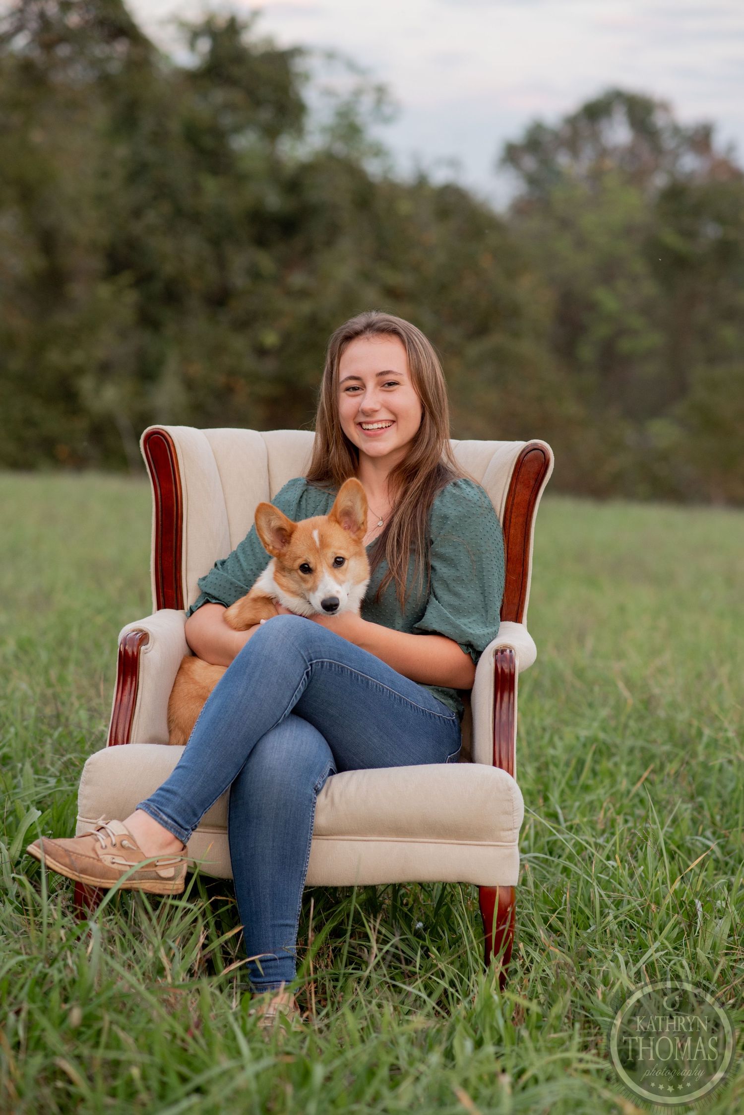Casual portrait of someone sitting in a beige armchair with a Corgi dog in their lap, set in an outdoor field.