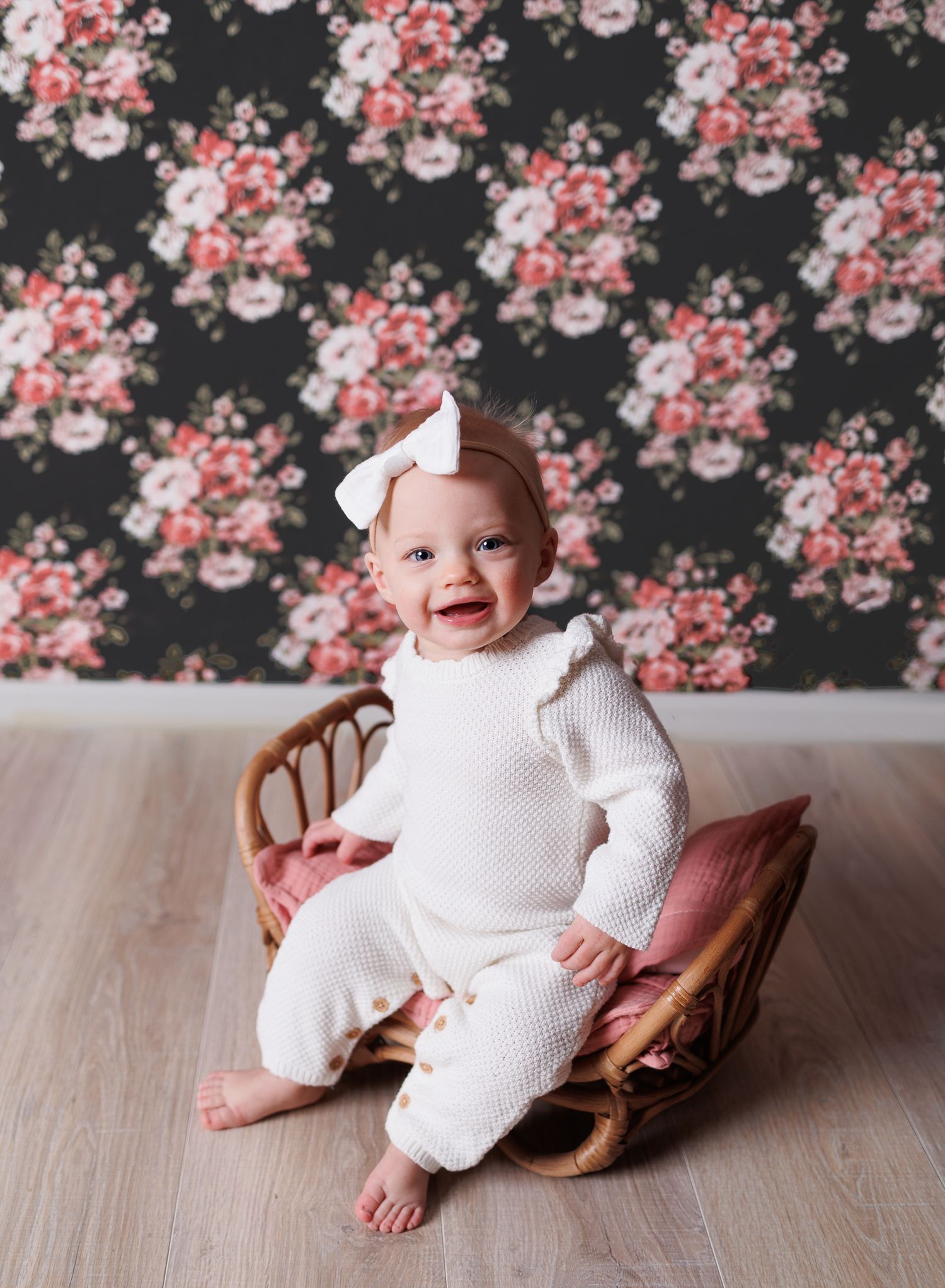Baby in white knit outfit sits on vintage chair against floral wallpaper.