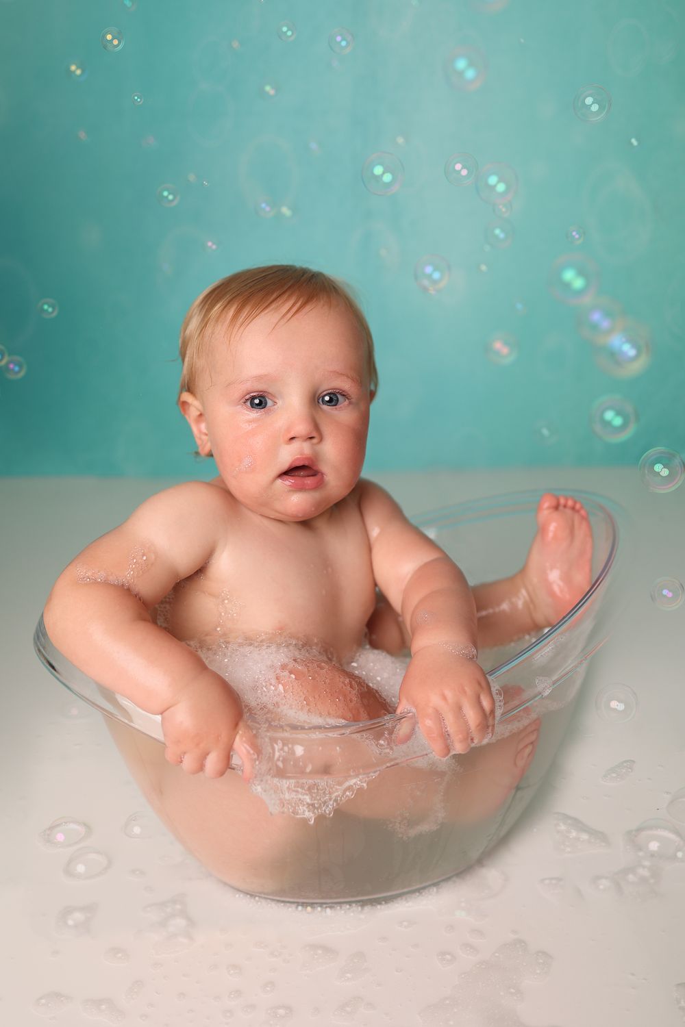 Baby sits in clear bowl with soap bubbles floating against turquoise background.