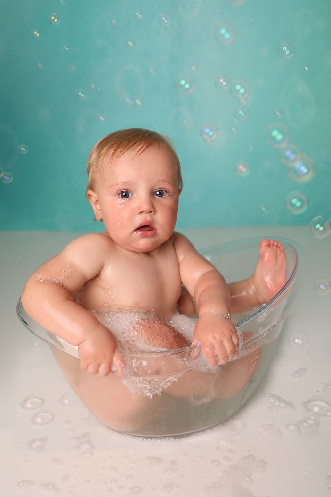 Baby sits in clear bowl with soap bubbles floating against turquoise background.