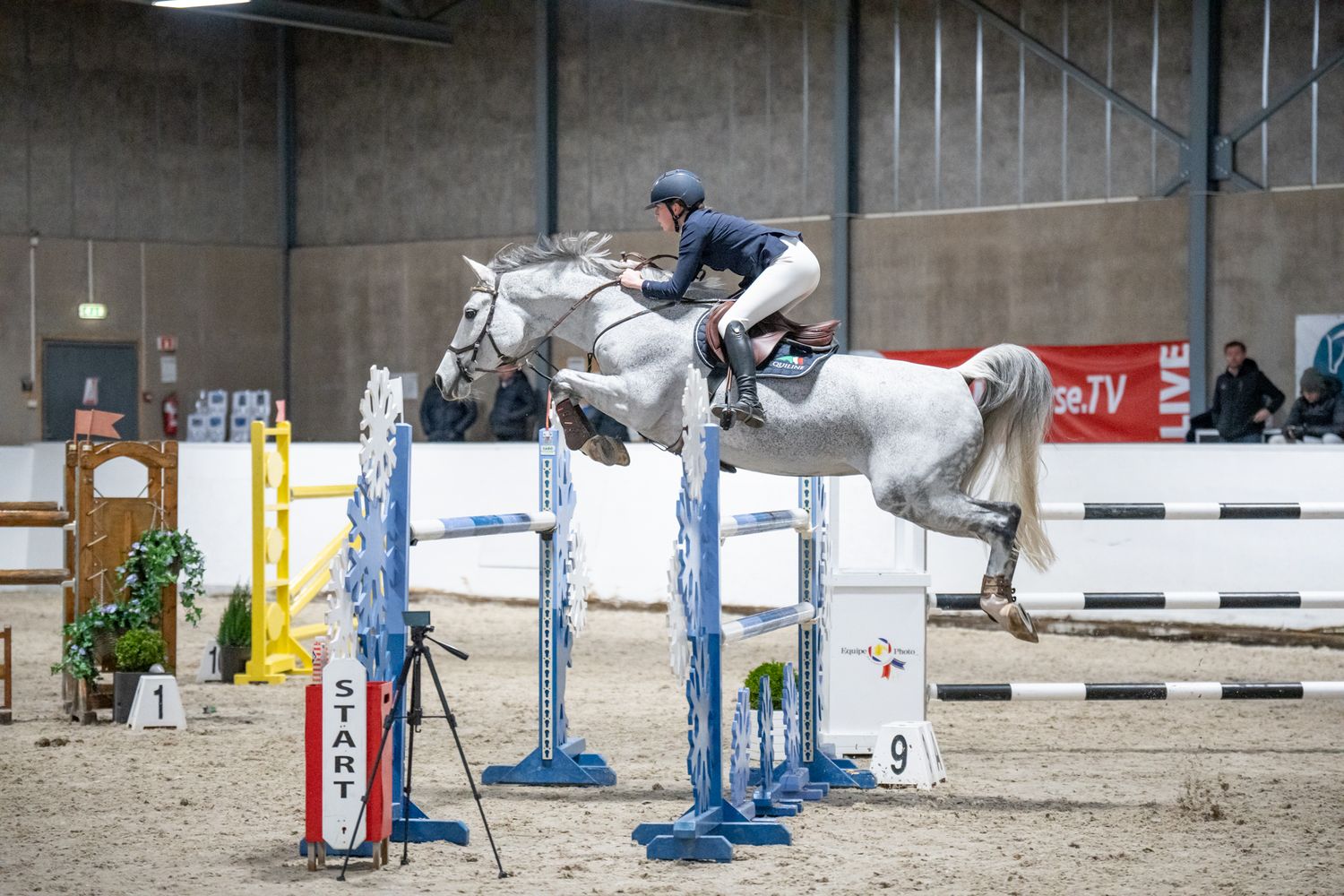 A grey horse and rider jumping over blue and white obstacles in a competition arena.