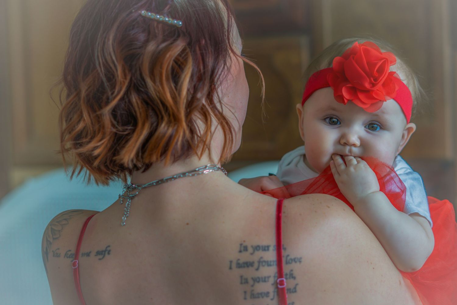 Tattooed mother holds baby girl with red flower headband, viewed from behind showing back tattoos.