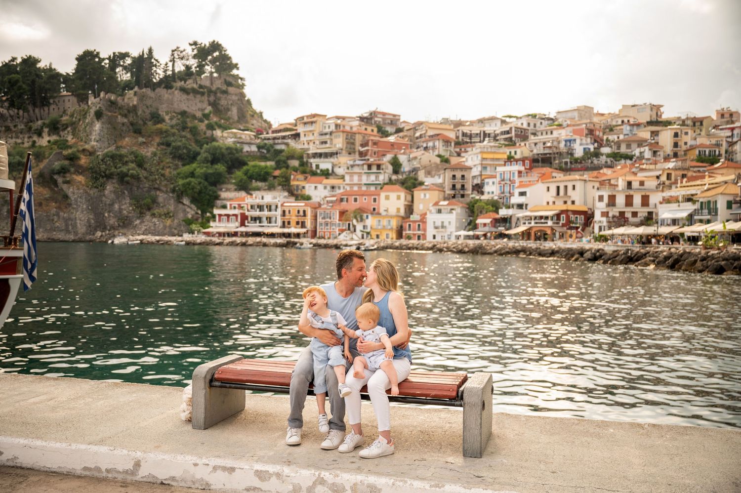 A family sits on a bench overlooking a picturesque Mediterranean coastal town with colorful buildings.