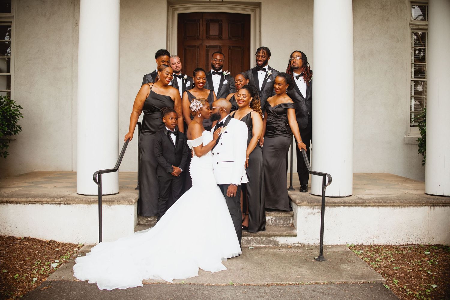 Wedding party in black formal attire poses with bride and groom in white on steps of a white colonial building.