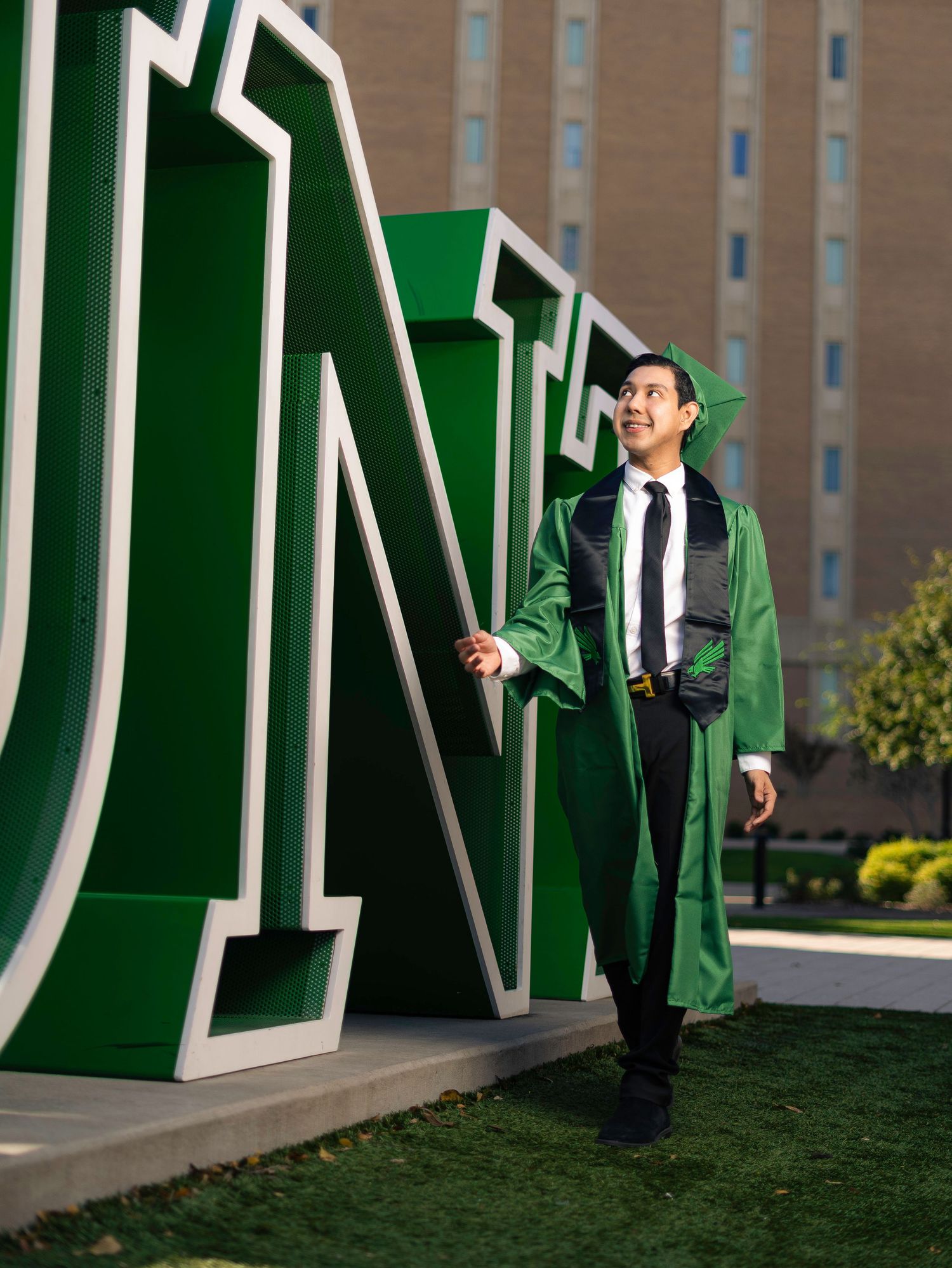Graduate poses beside large green UNT letters on campus.