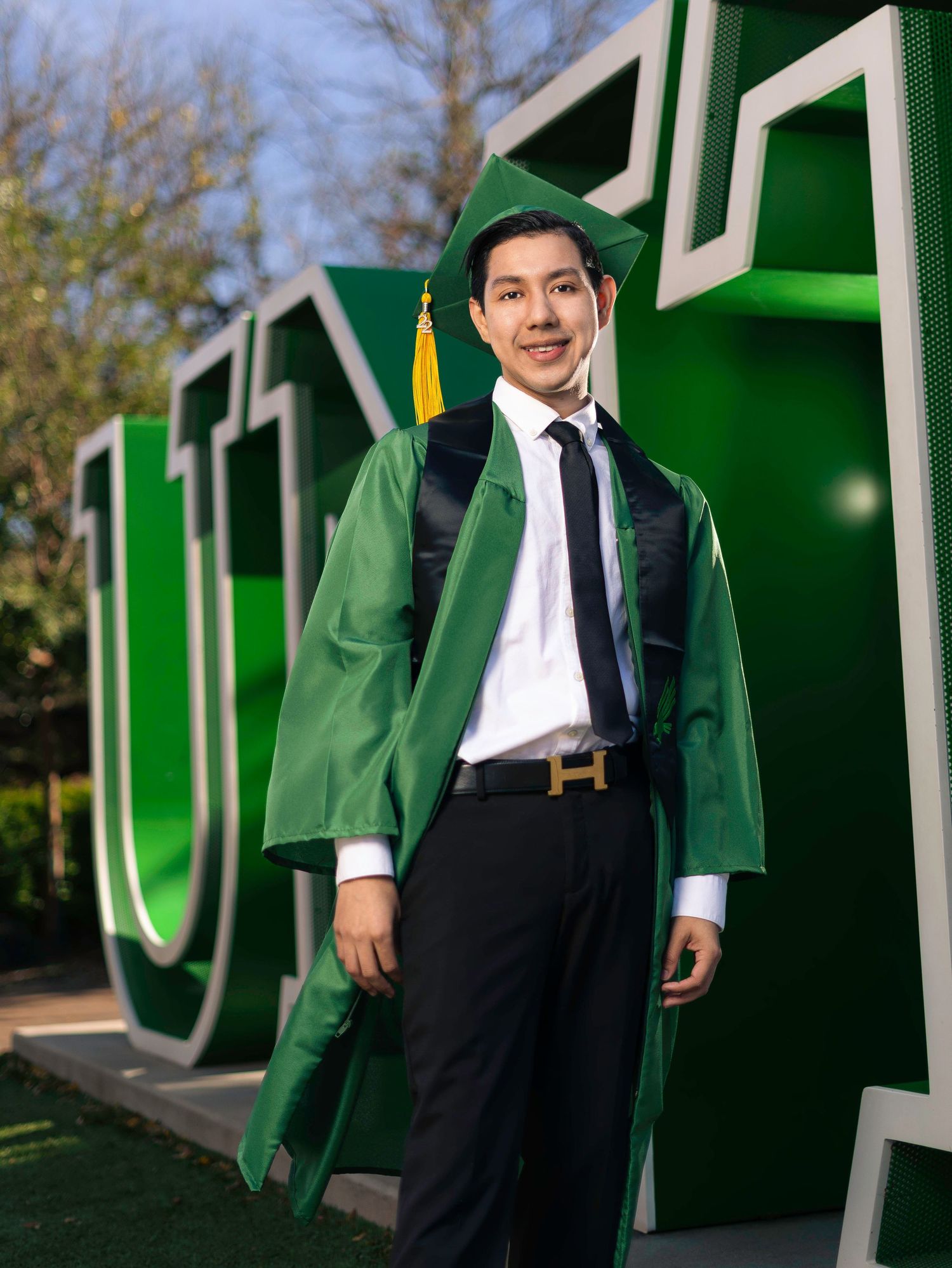 Graduate in green regalia stands proudly in front of large UNT lettering.