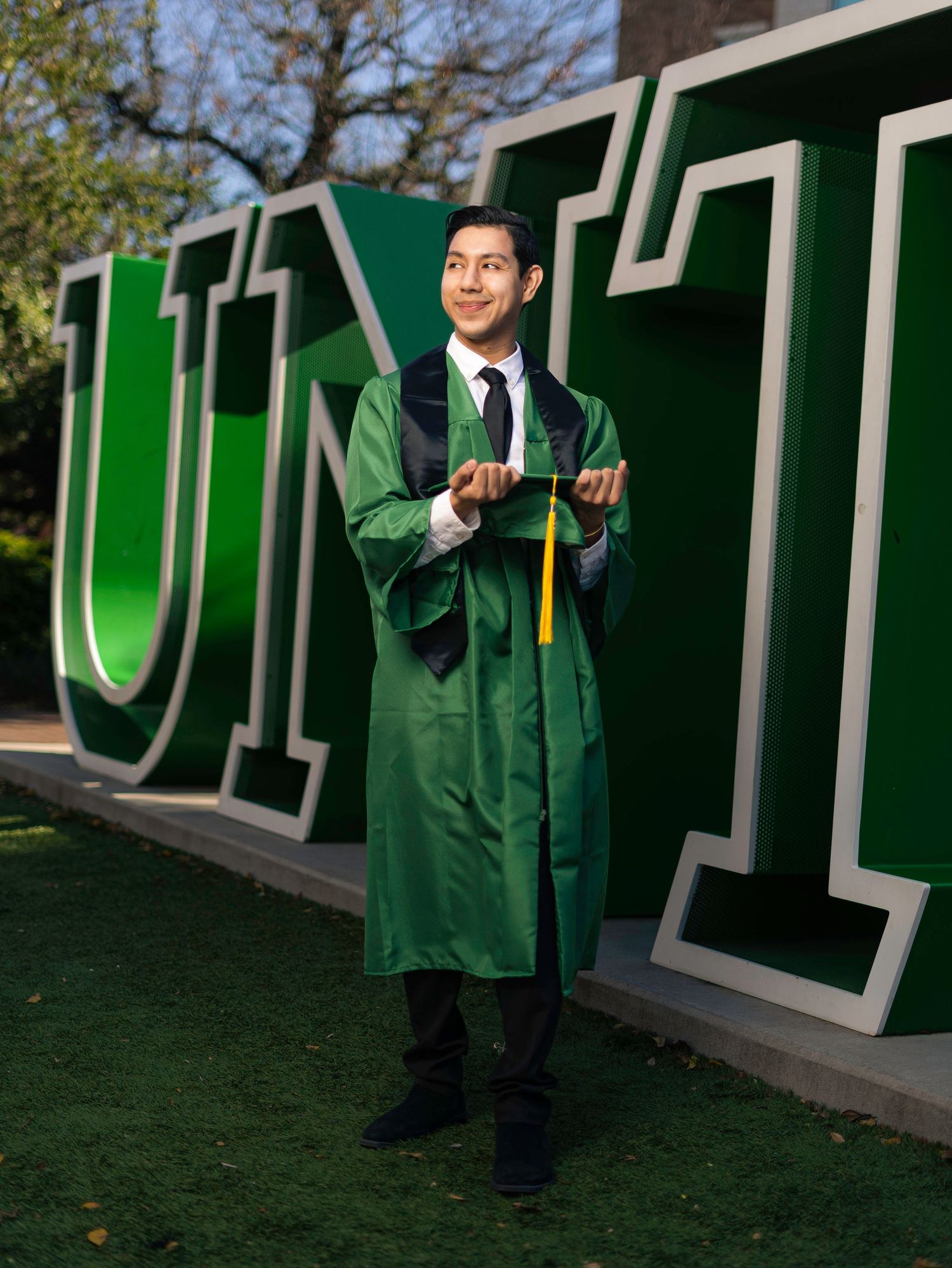 Graduate in green regalia poses in front of large university letters outdoors.