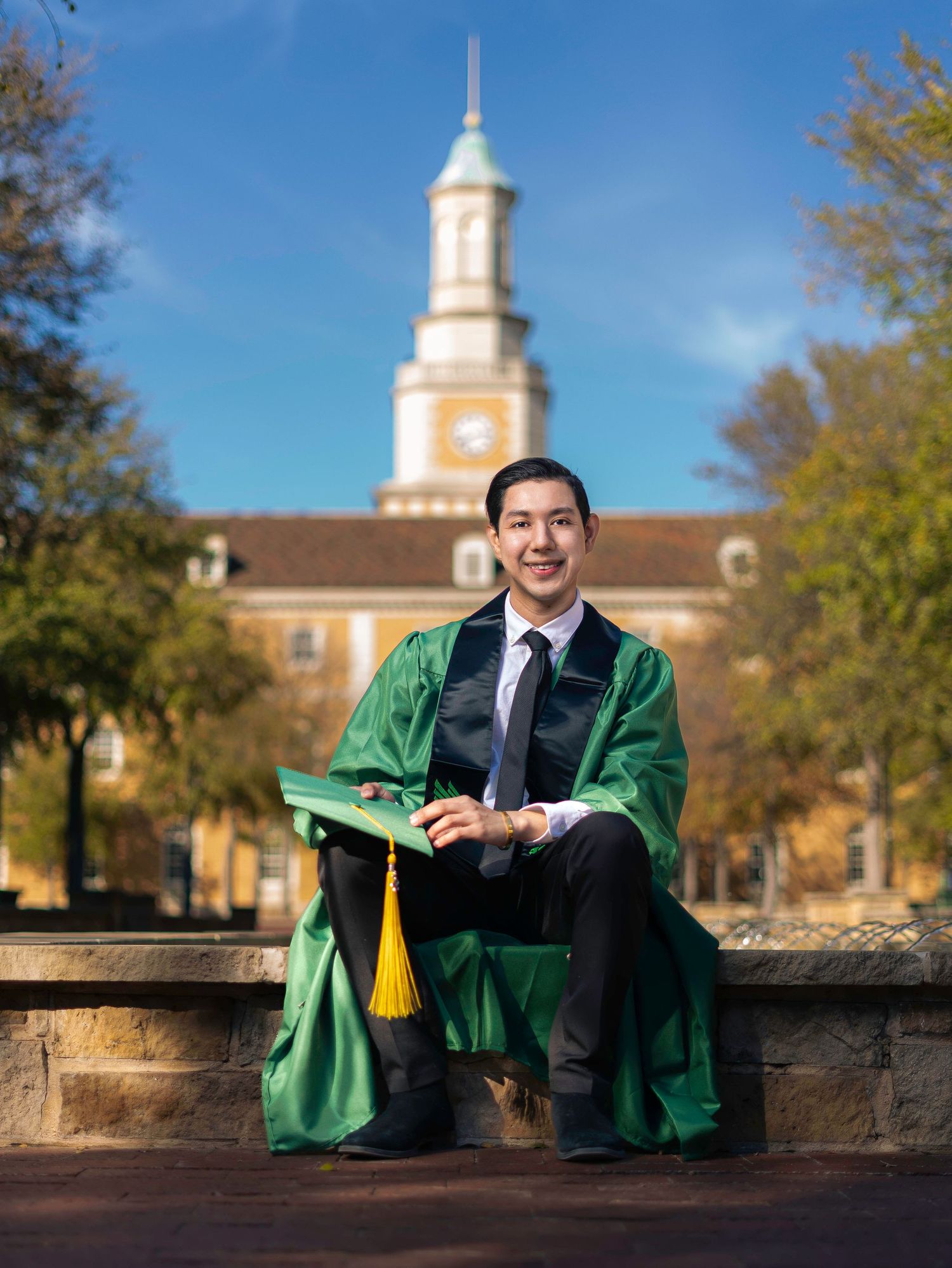 Graduate in green and black cap and gown poses on campus with historic clock tower in background.