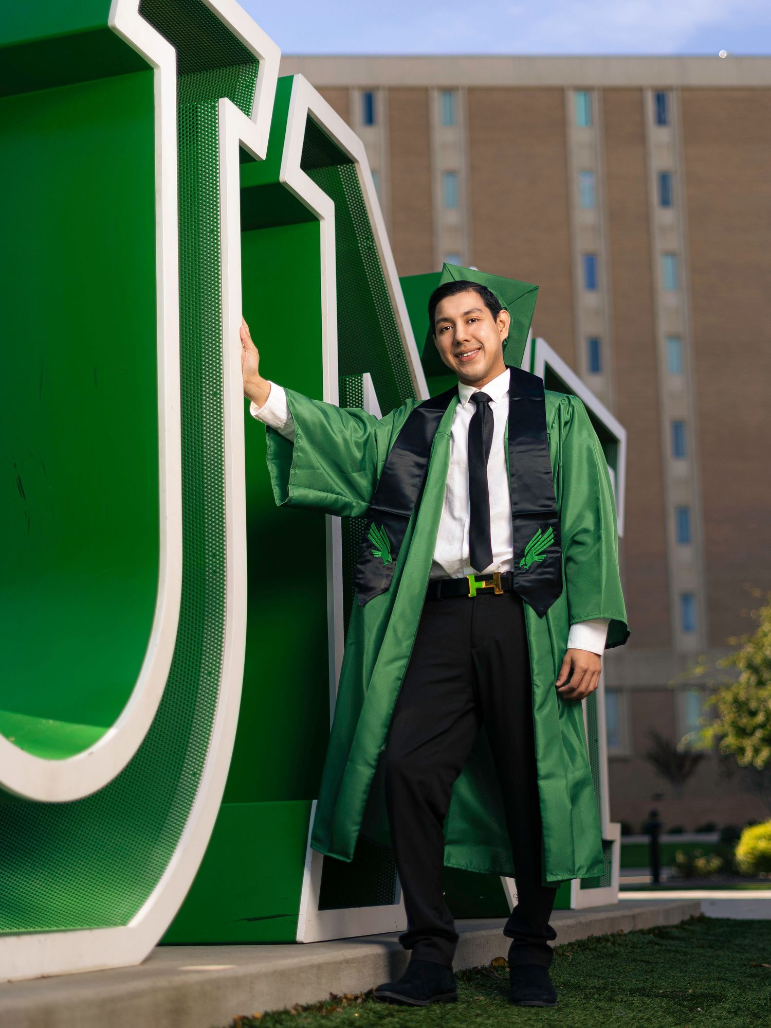 Graduate in green commencement gown poses beside large university letters.