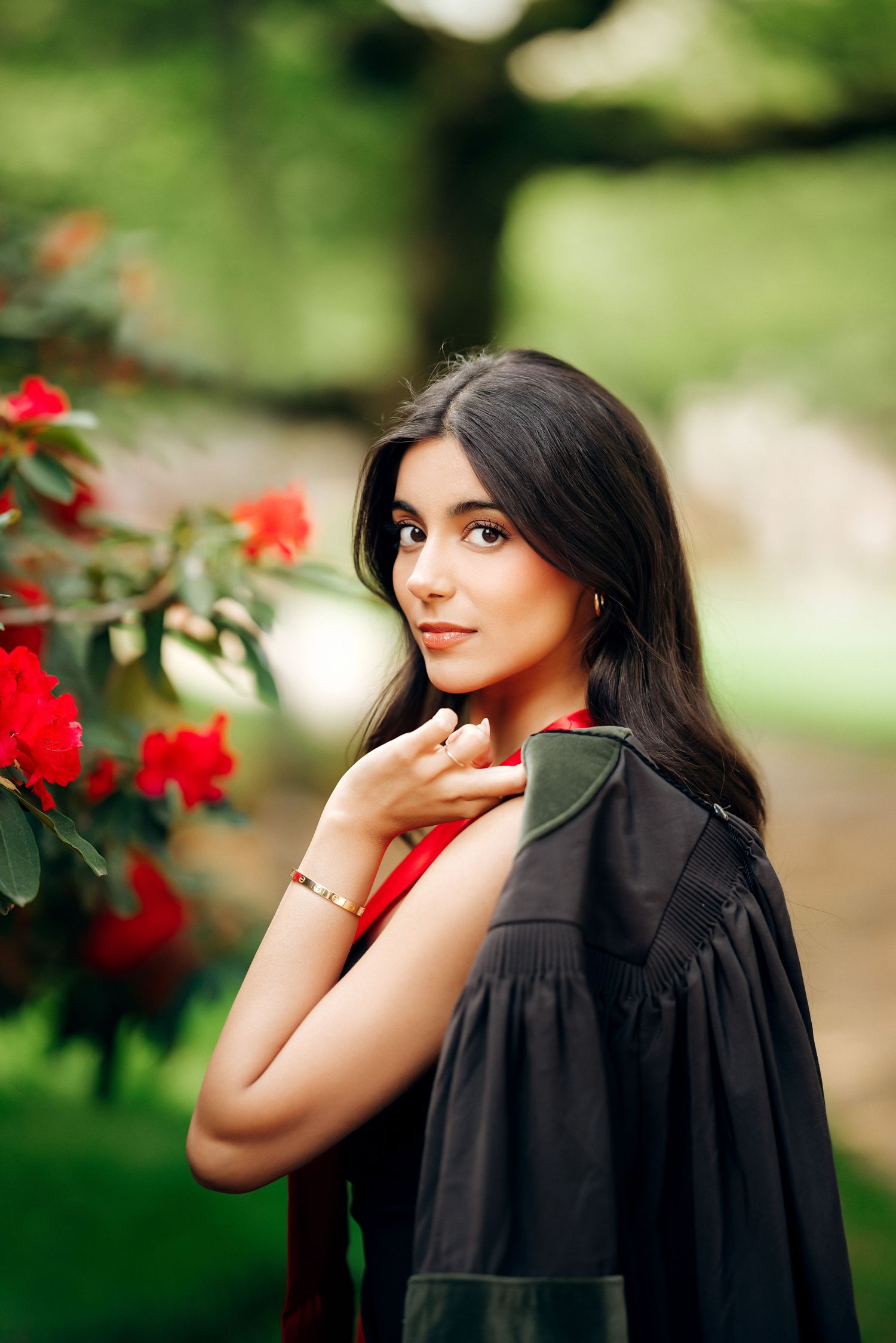 A woman in a black dress poses elegantly next to blooming red roses in a garden.