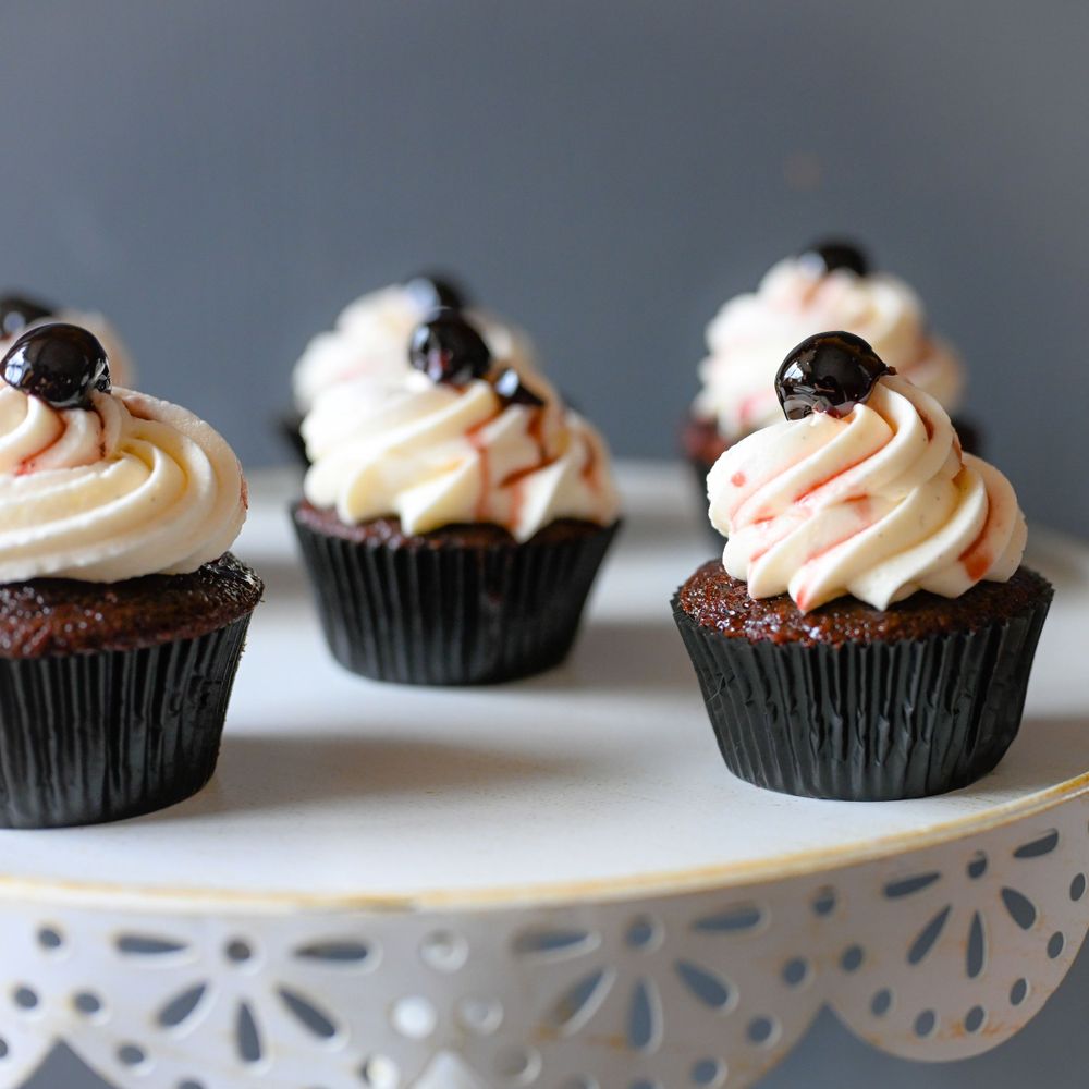 Chocolate cupcakes topped with swirled frosting and dark chocolate cherries on a decorative white stand.