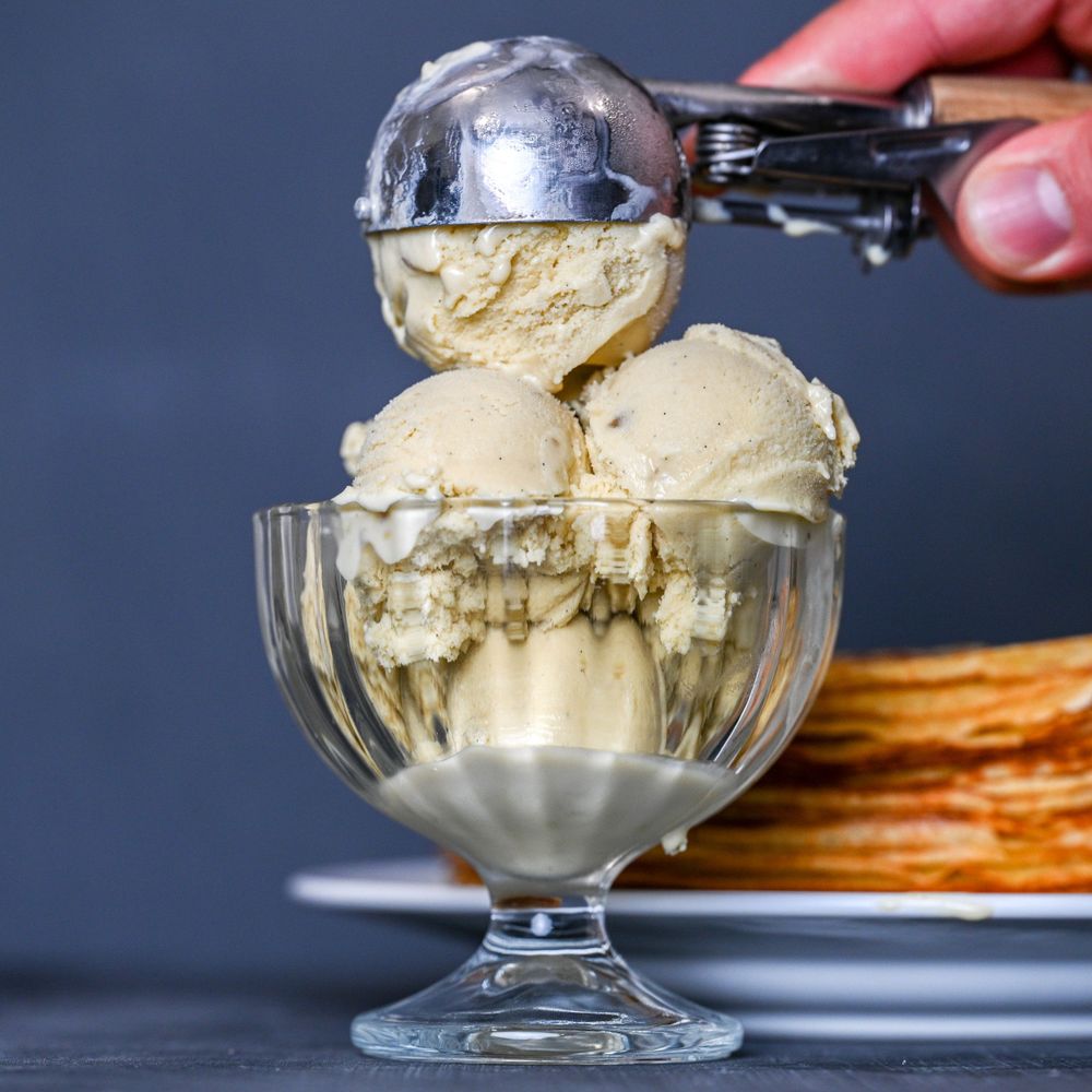 Ice cream being scooped into a glass dessert bowl with pancakes visible in the background.