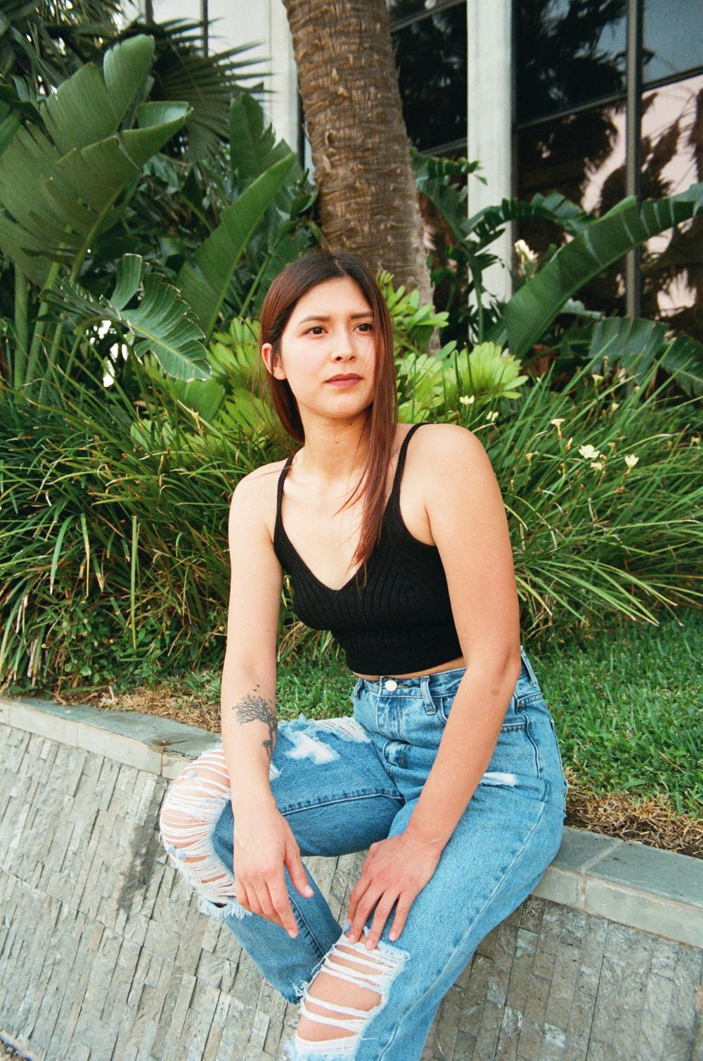 Young woman in black crop top and ripped jeans sitting outdoors near tropical plants.