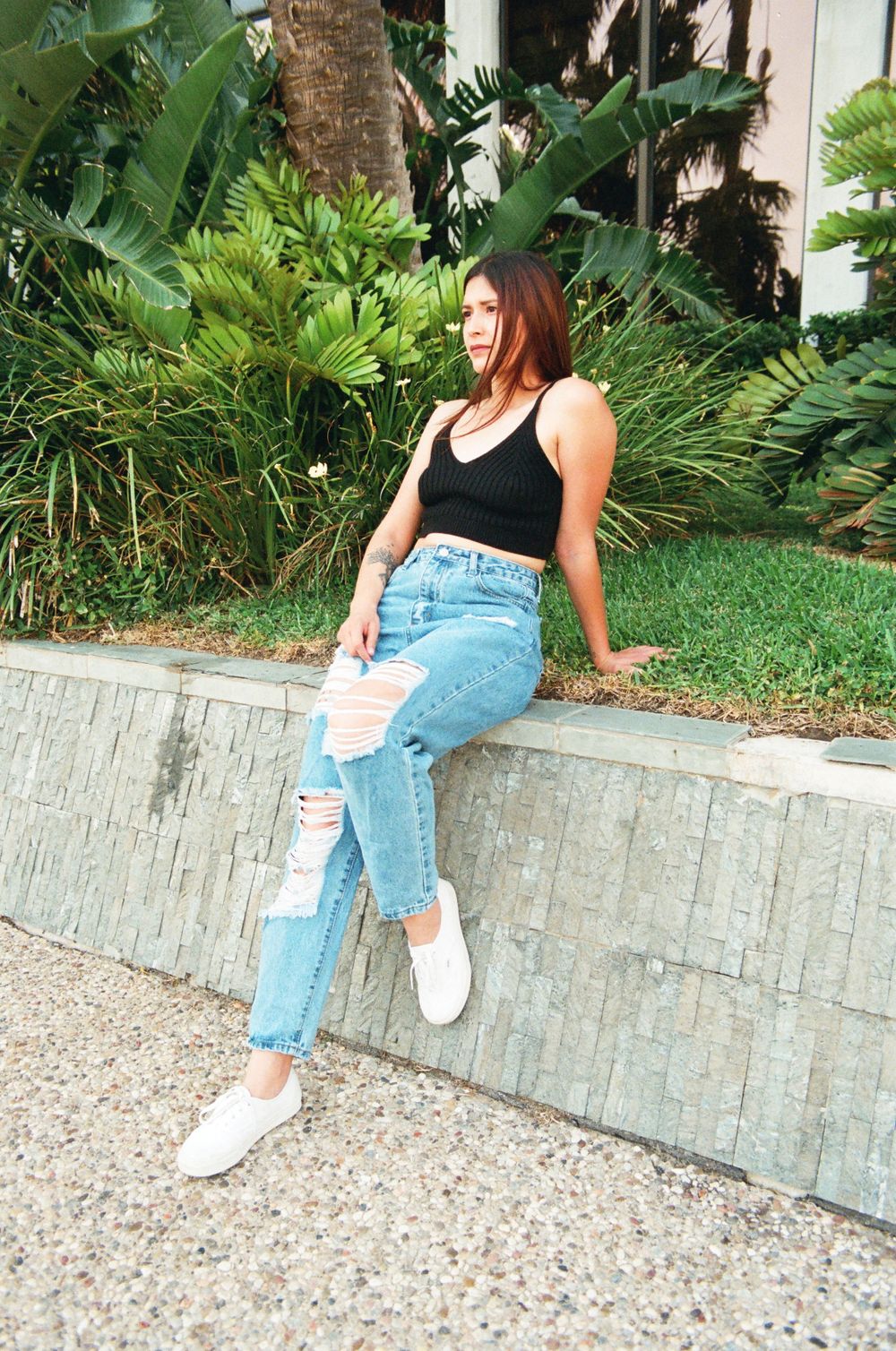 Young woman in black crop top and ripped jeans leaning on stone ledge with tropical plants behind.