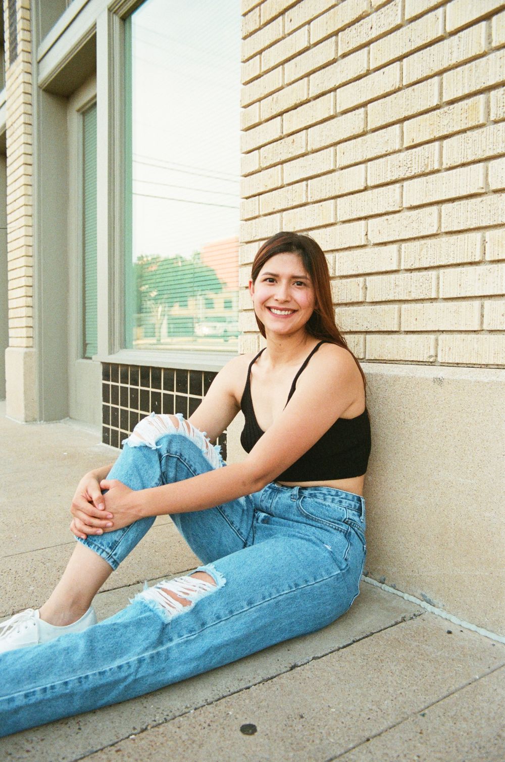Young woman in black crop top and ripped jeans sitting against brick wall, smiling warmly.
