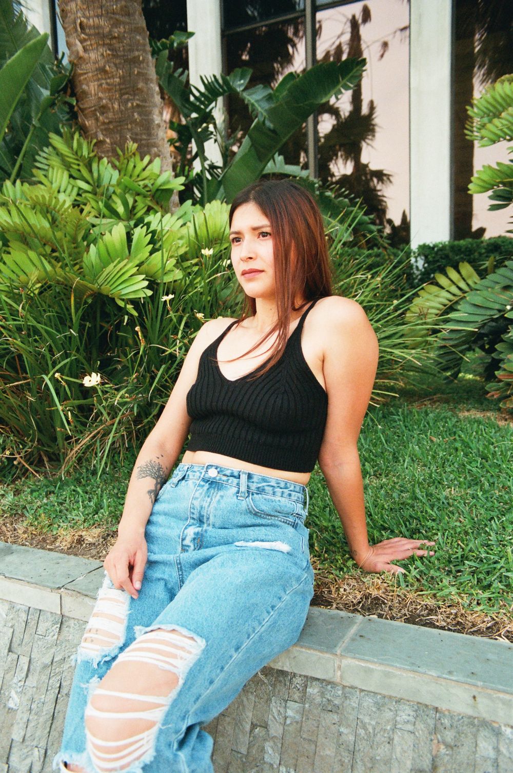 Young woman in black crop top and distressed jeans sitting on ledge with lush green plants behind.