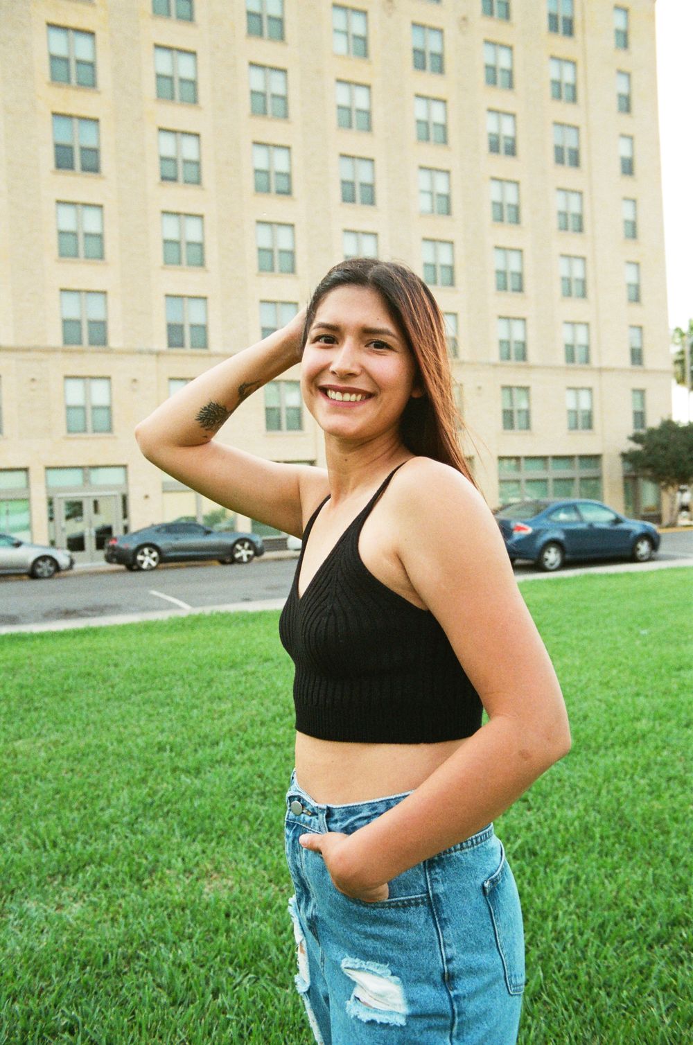 Smiling young woman in black crop top and ripped jeans posing on green lawn near building.