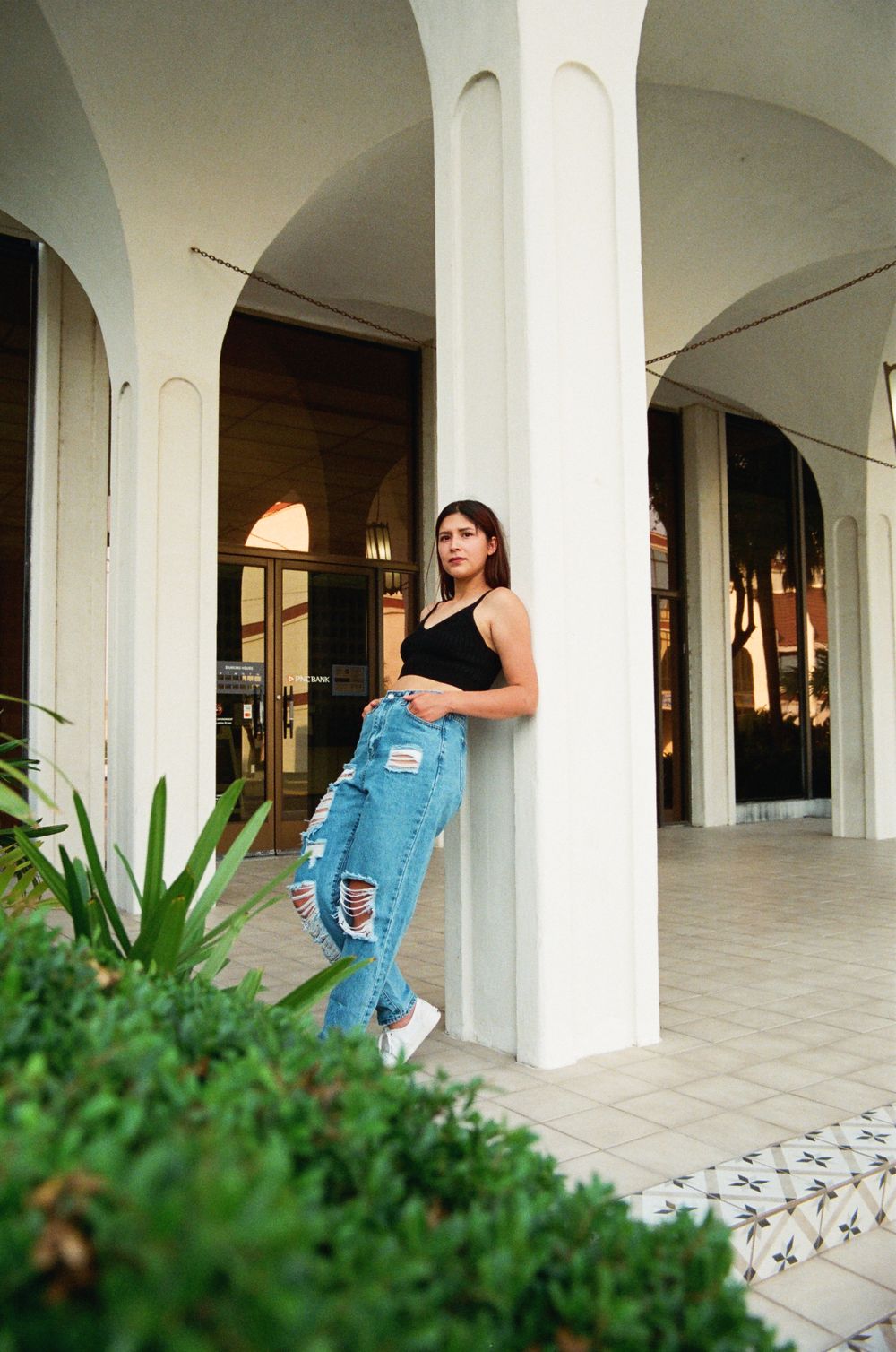 Stylish woman in black tank top and distressed jeans leaning against white column in arched building.