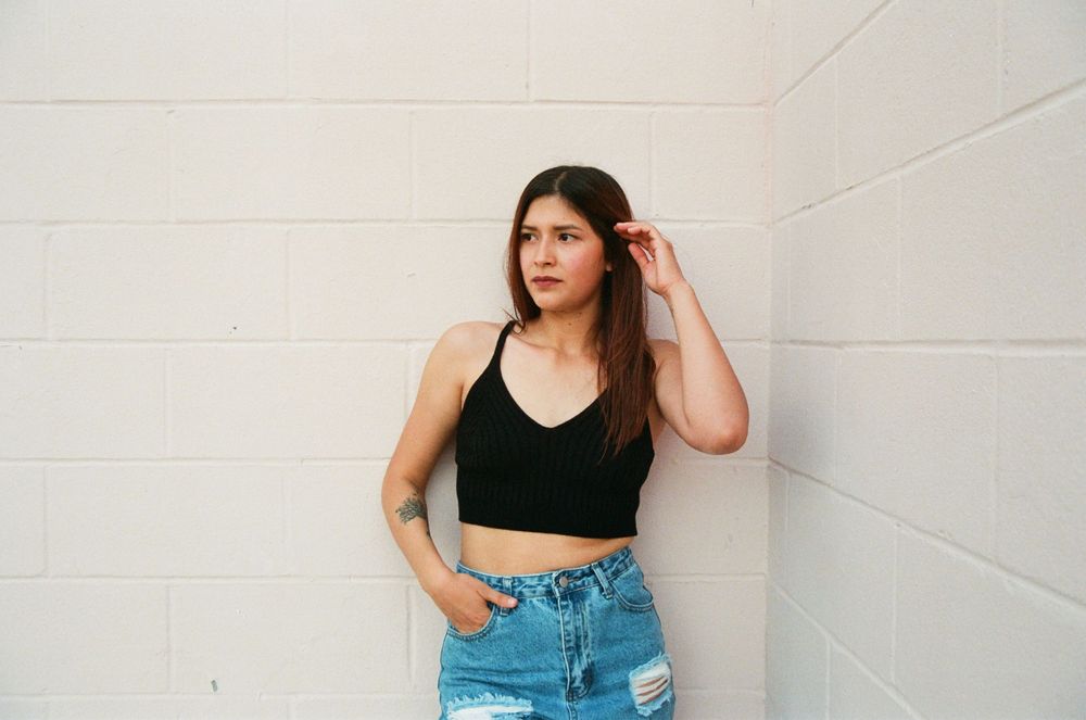 Young woman in black crop top and ripped jeans poses against white brick wall.