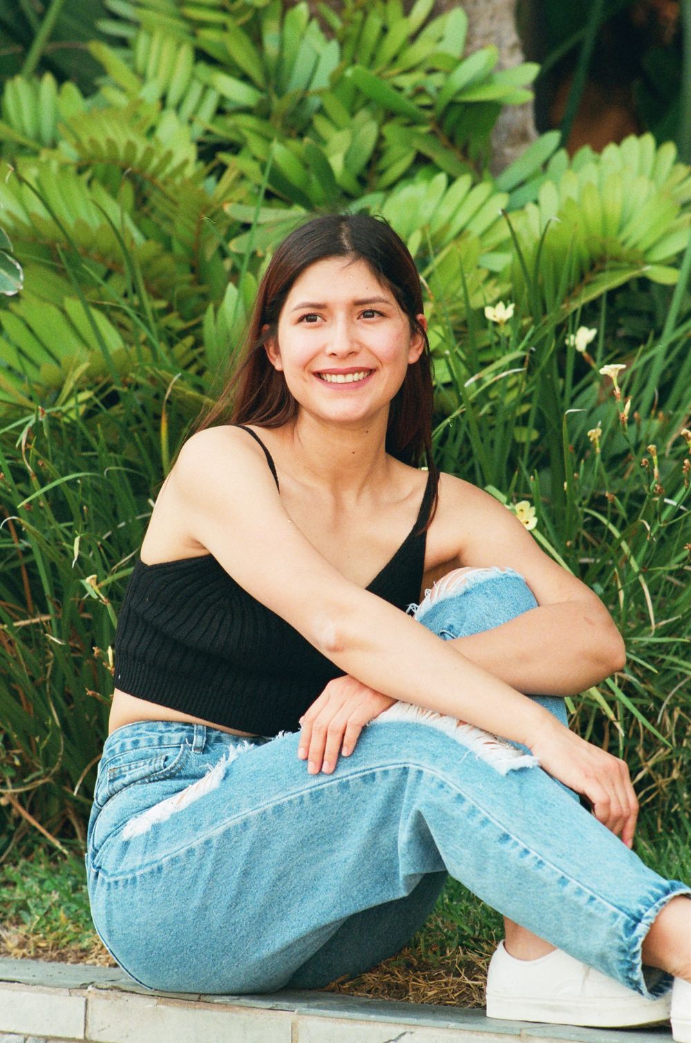 Smiling young woman in black crop top and ripped jeans sitting on ground near lush green plants.