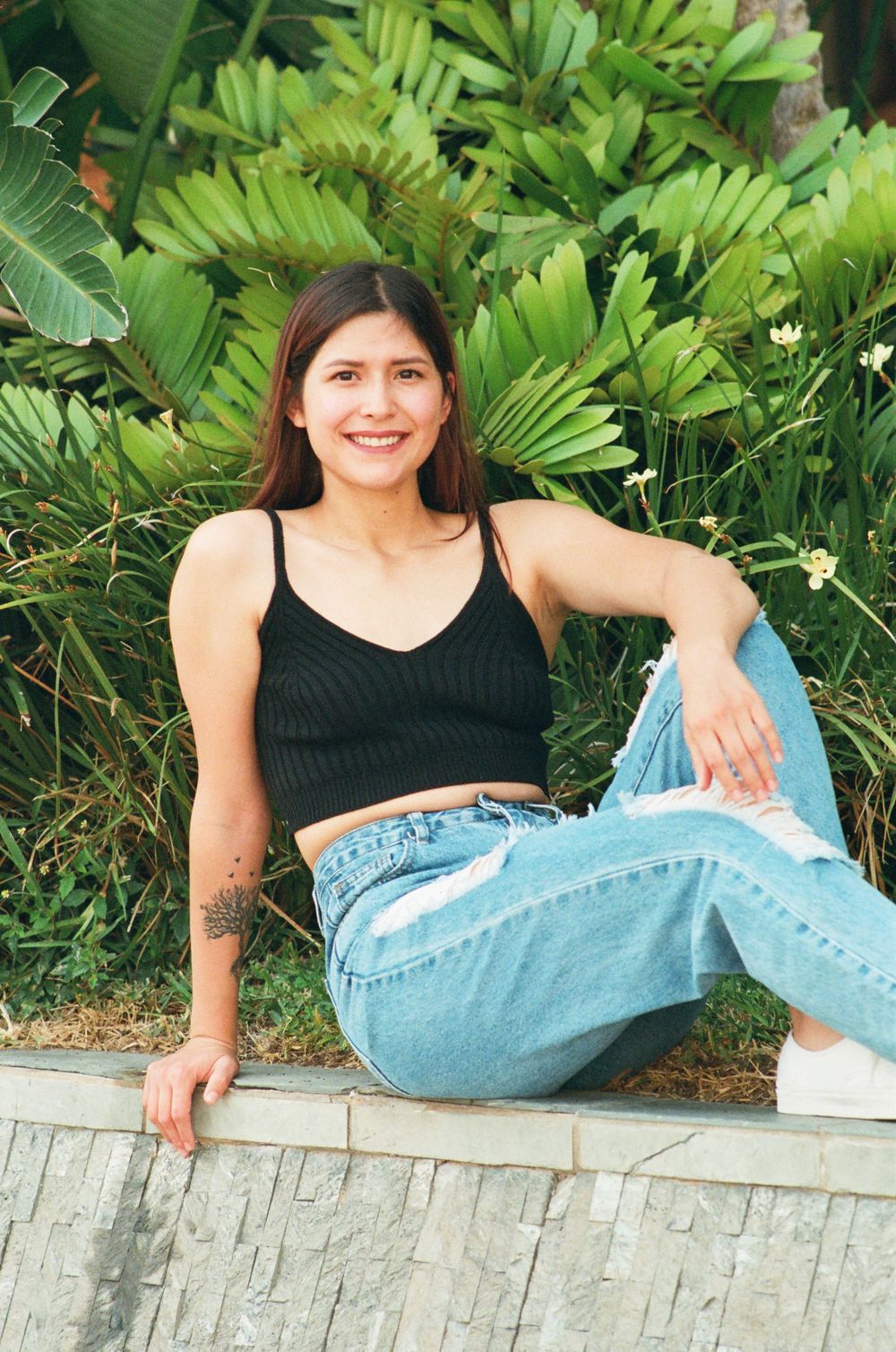 Happy young woman in black crop top and distressed jeans sitting outdoors against tropical greenery.