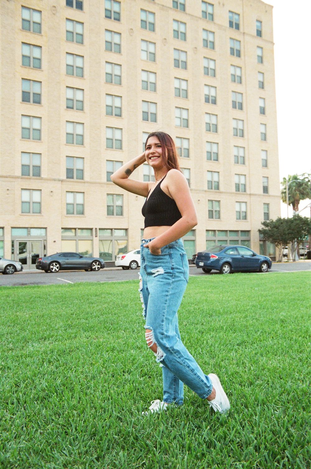 Young woman in black crop top and ripped jeans walking on green grass near tall building.