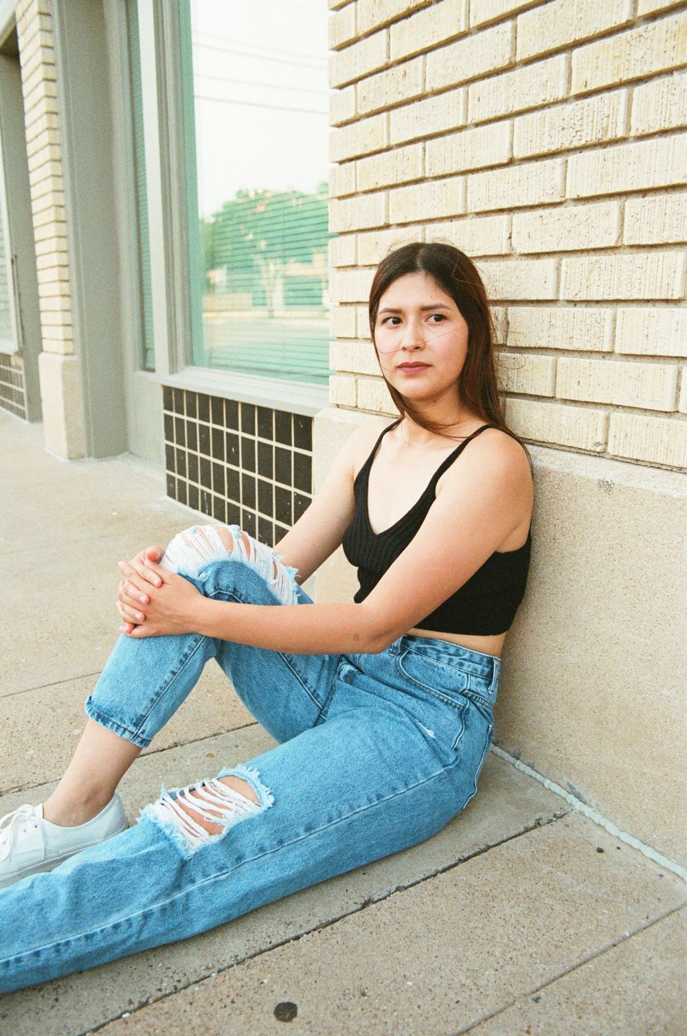 Young woman in black crop top and distressed jeans sitting against brick wall with serious expression.