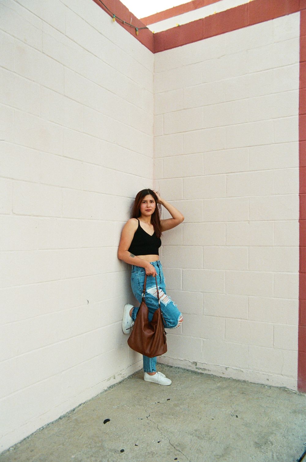 Young woman in black tank and ripped jeans poses in brick corner holding brown bag.