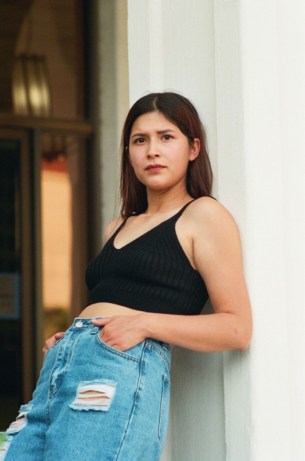 Young woman in black ribbed crop top and ripped jeans leaning against white wall outdoors.