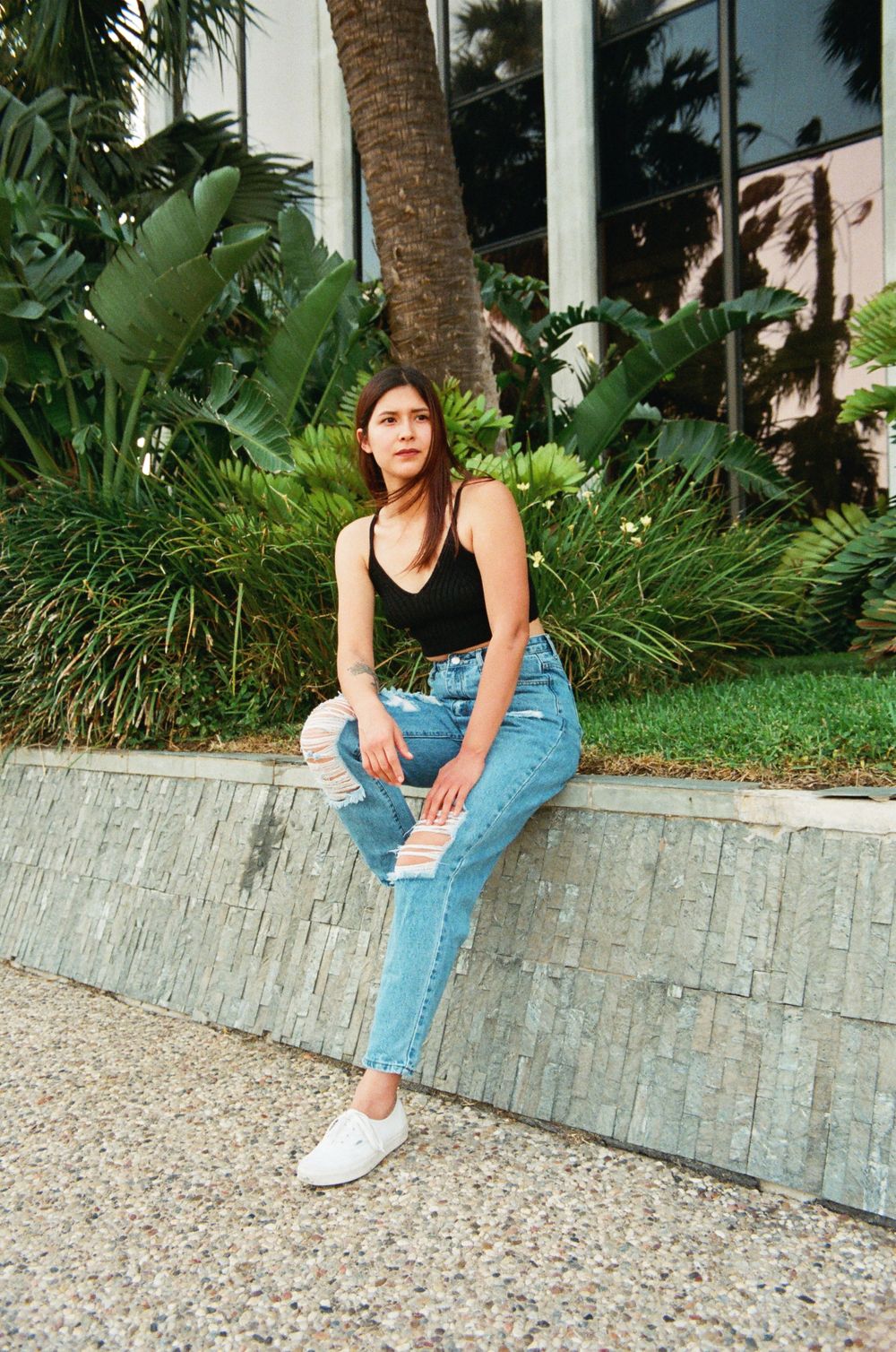 Young woman in black crop top and light ripped jeans sitting on stone ledge with tropical background.