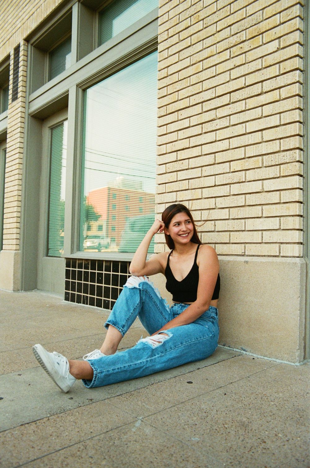 Young woman in black tank and ripped jeans sitting against brick wall outdoors, smiling at camera.
