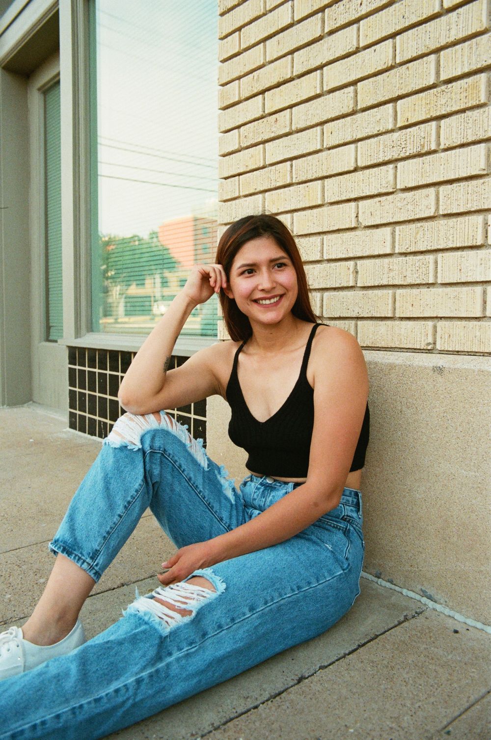 Smiling young woman in black crop top and distressed jeans sitting against brick building wall.