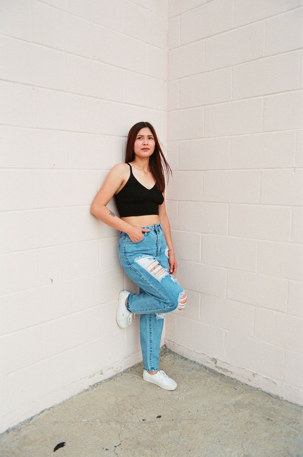 Young woman in black crop top and ripped jeans leans against white brick wall corner.