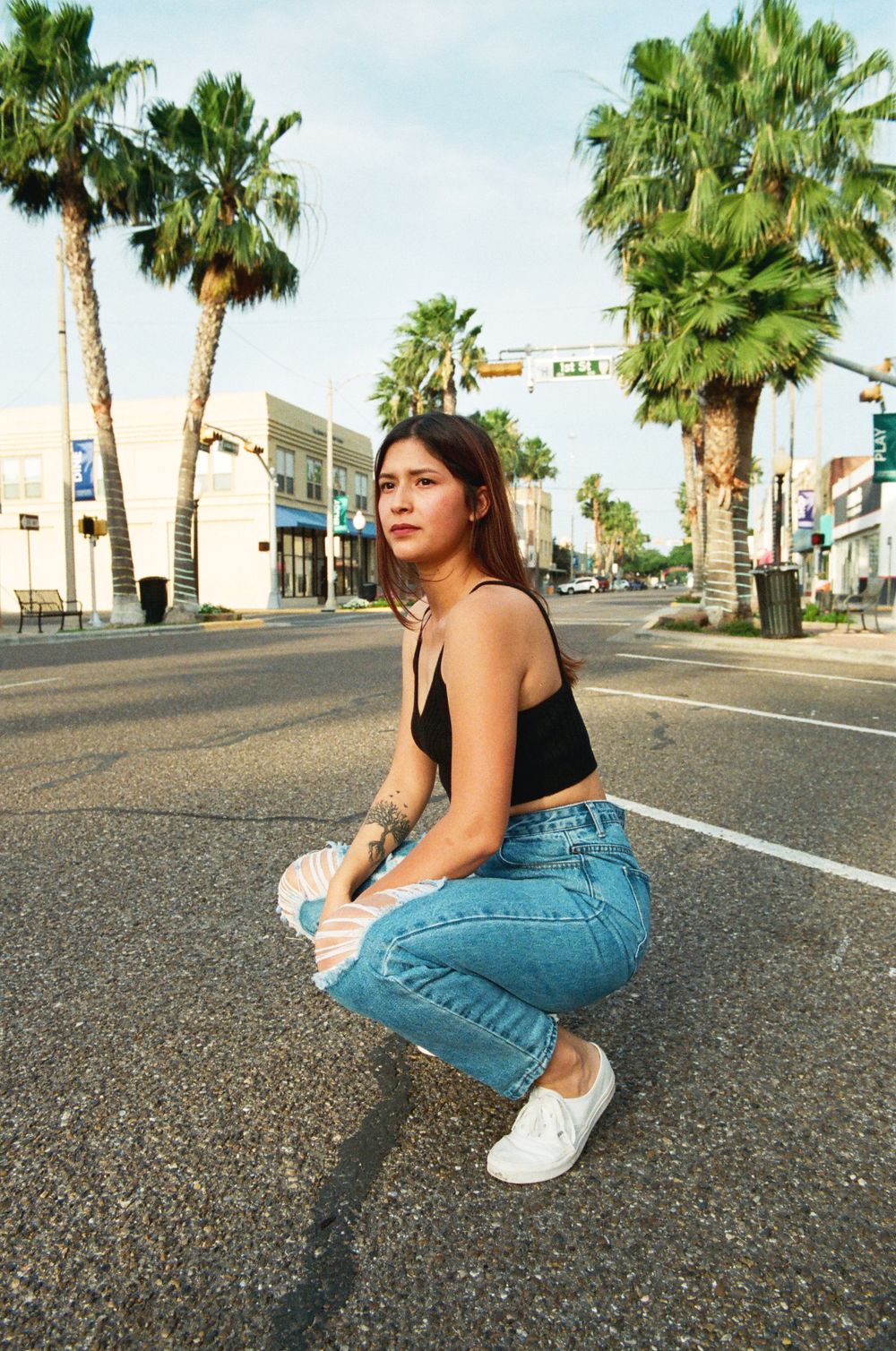 Young woman in black crop top and jeans crouching on urban street with palm trees behind her.