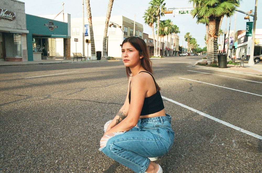 Young woman in black crop top and jeans crouching on empty street lined with palm trees.