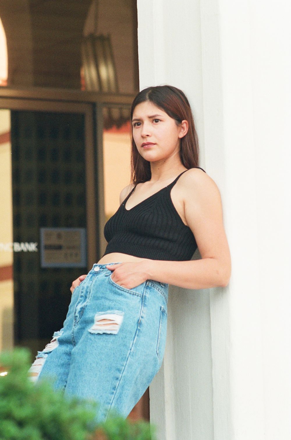 Woman in black spaghetti strap top and distressed denim jeans posing against white pillar outdoors.