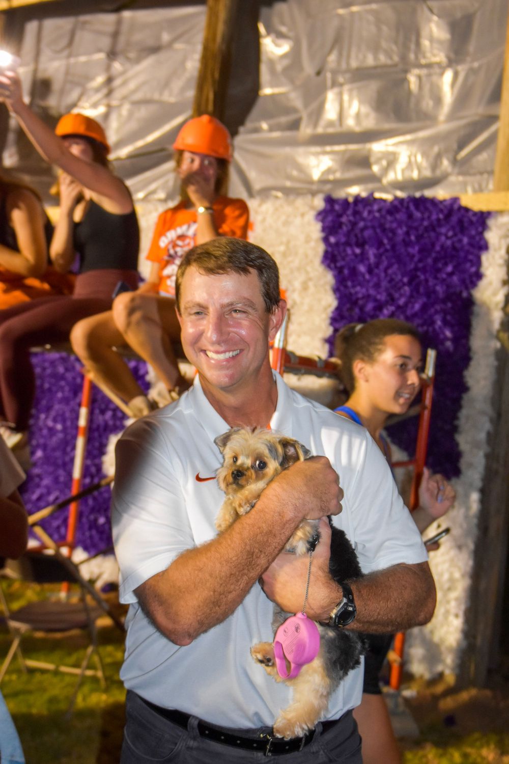 Person in white polo shirt holding a small dog at outdoor event with purple decorations.