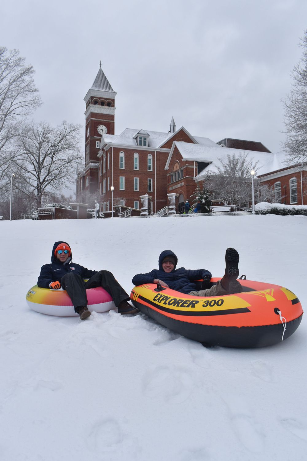 Two people snow tubing down a hill in front of a brick church building in winter.