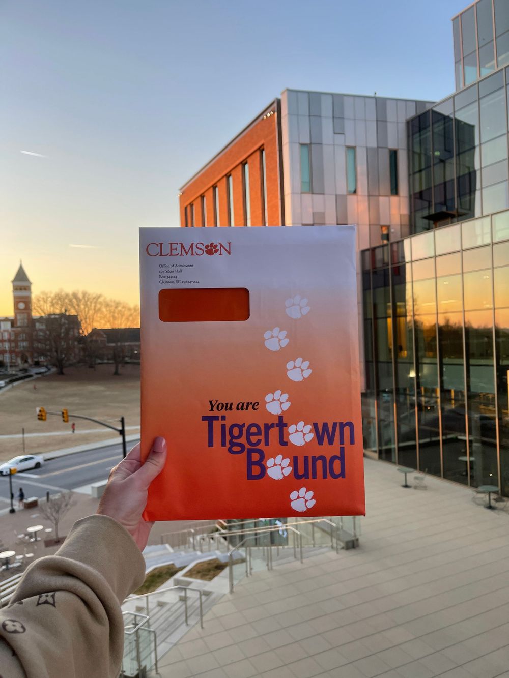Hand holding Clemson Tiger Bound acceptance letter against modern campus building at sunset.