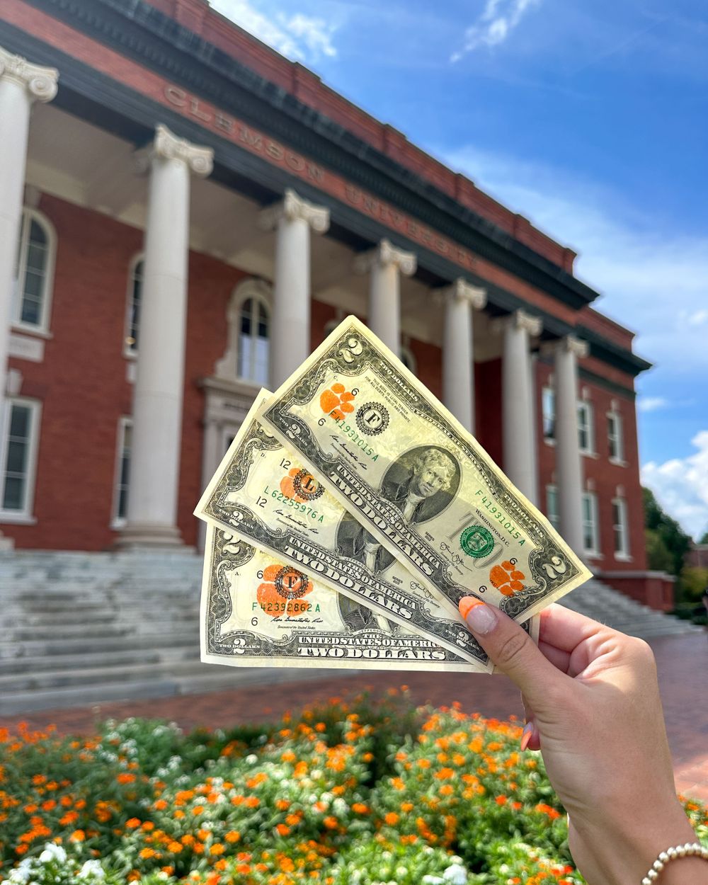 Hand holding decorated dollar bills in front of red brick university building with columns.
