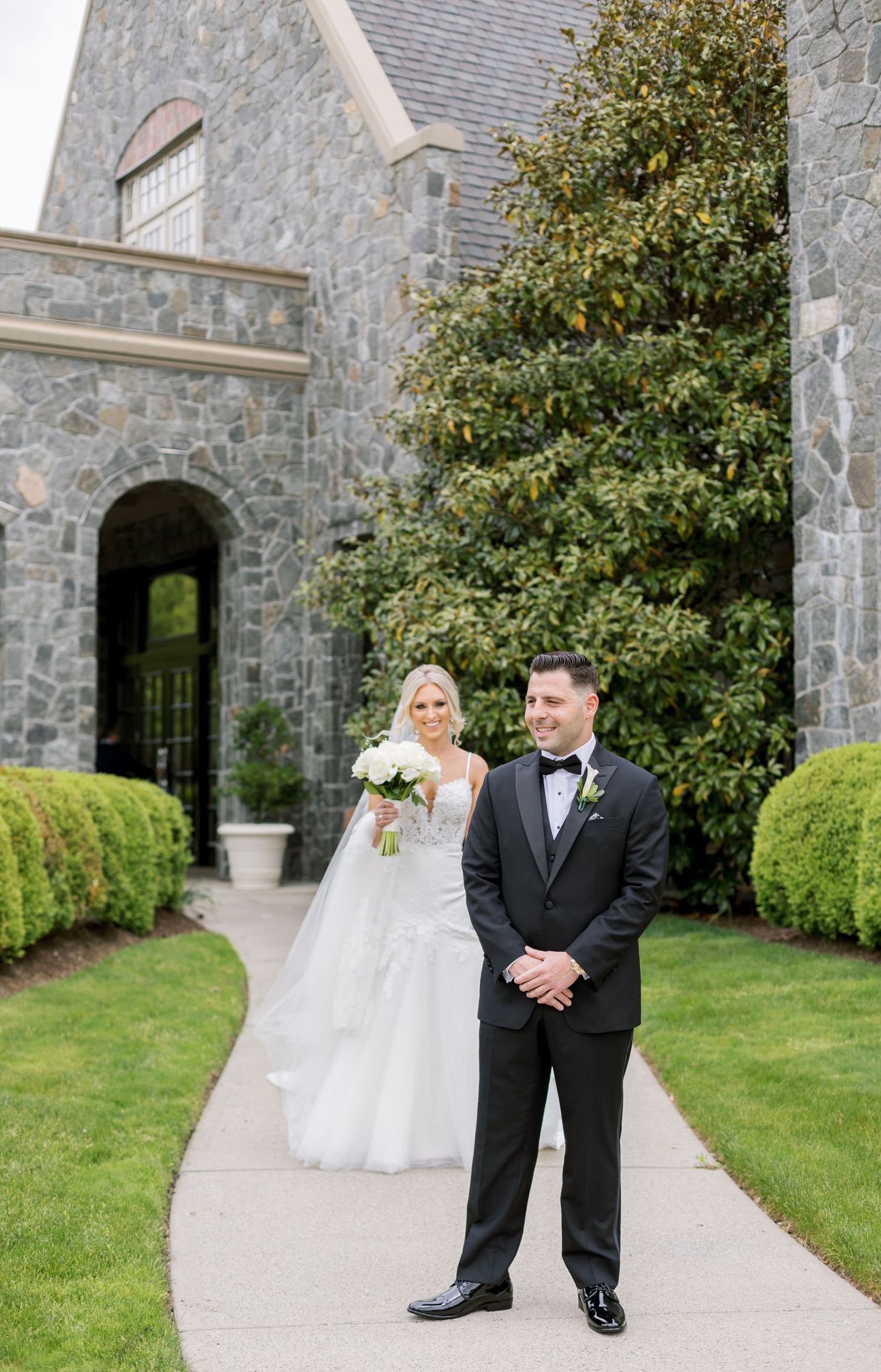 Wedding couple poses in front of stone church with beautiful landscaping.