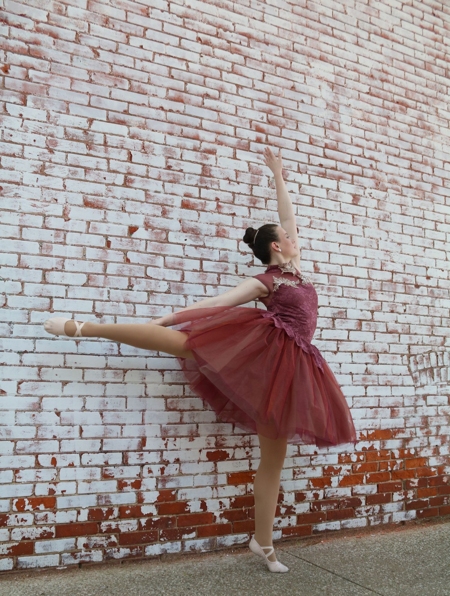 A graceful ballet dancer in a burgundy tulle dress poses against a white brick wall.