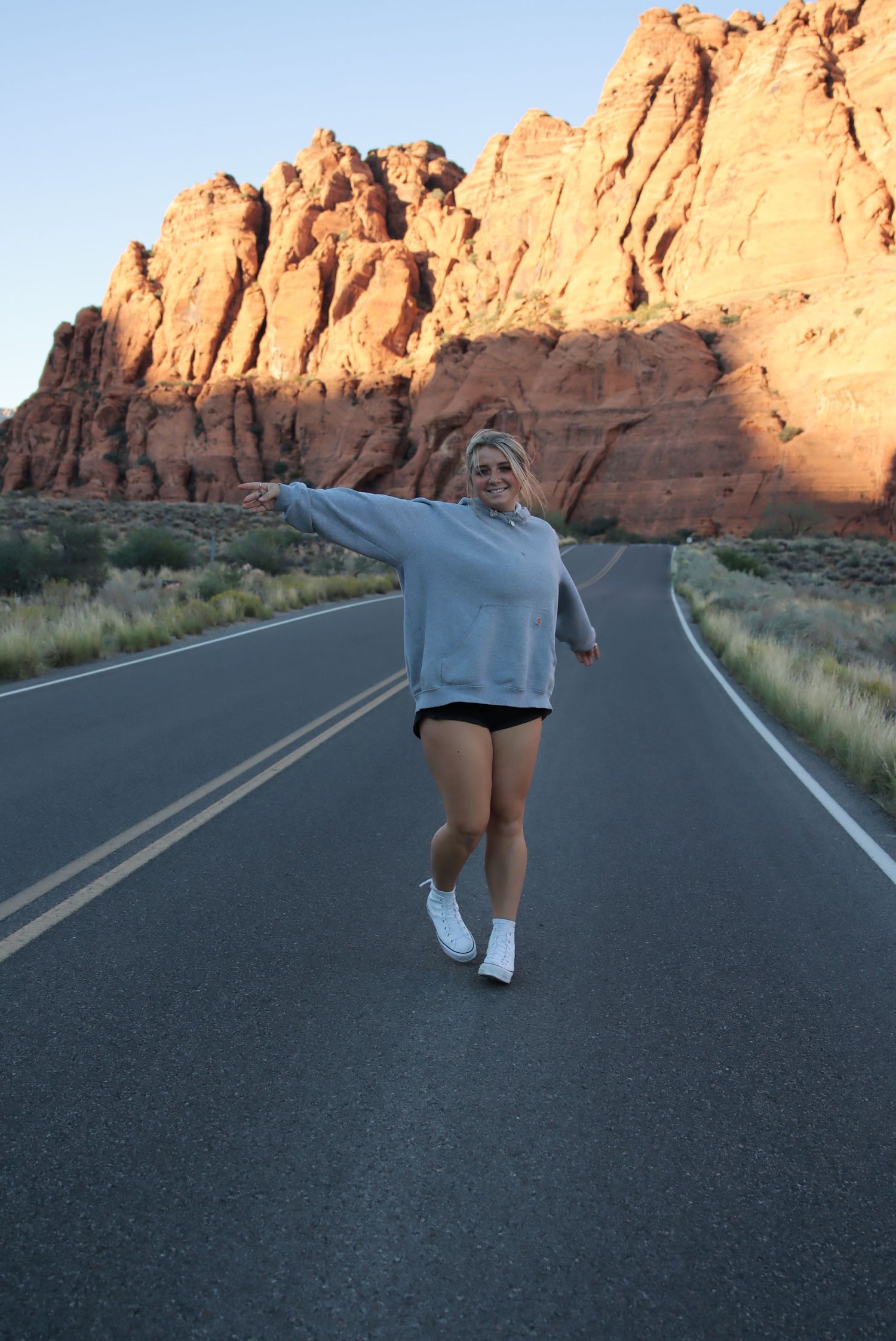 Cora Emilie Media Person in gray sweatshirt posing on empty road through red rock canyon.