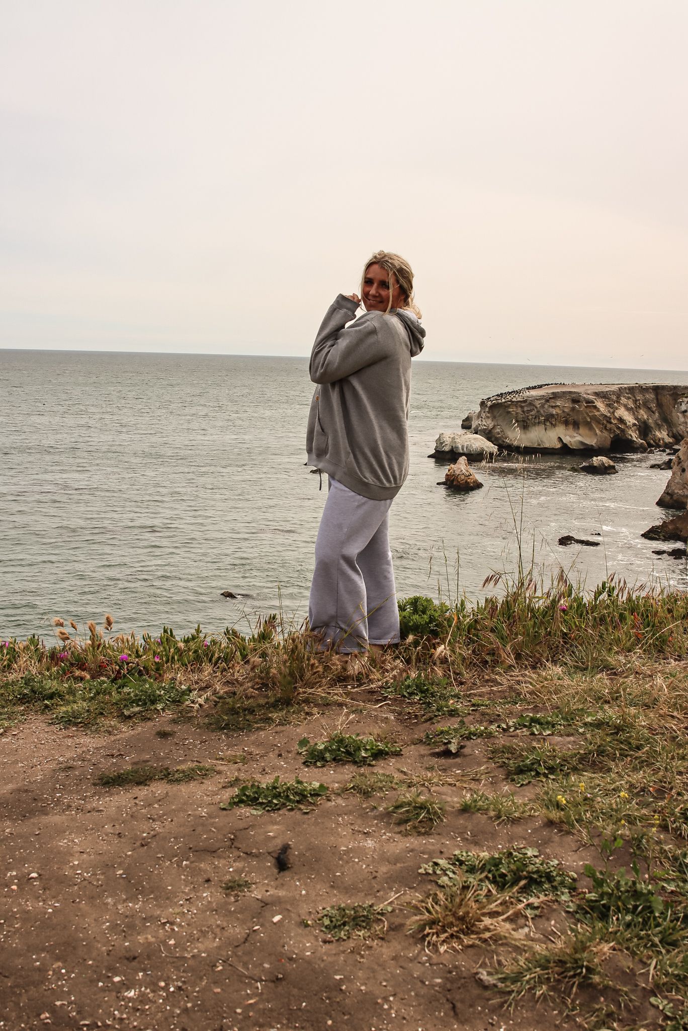 Cora Emilie Media Figure in gray sweater stands on coastal cliff overlooking rocky ocean shoreline on cloudy day.