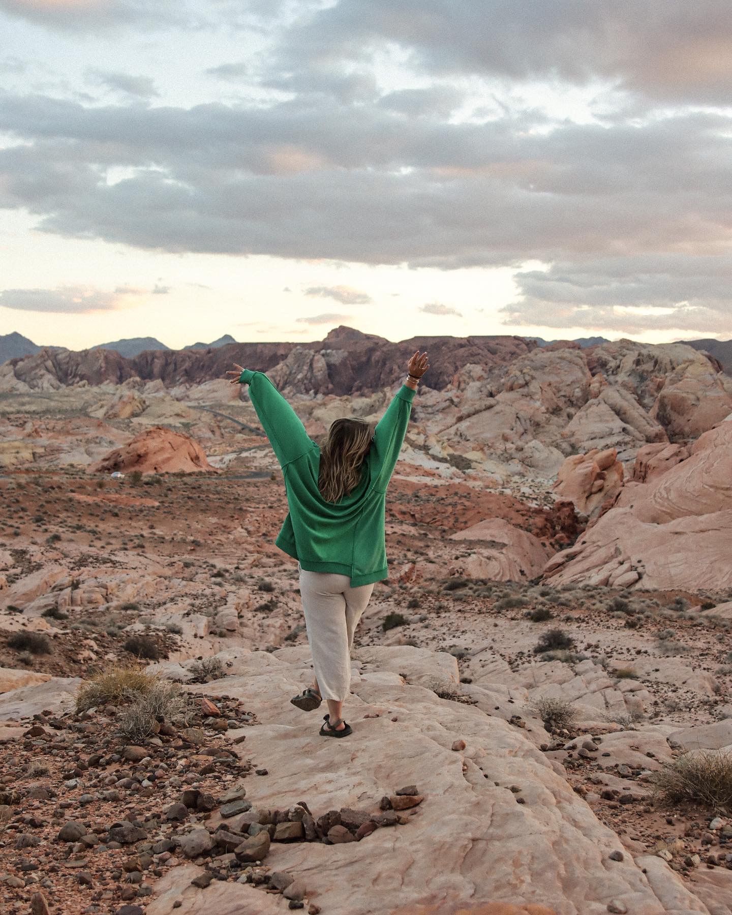 Cora Emilie Media Figure in green sweater raises arms triumphantly while standing on red rock formations at sunset.