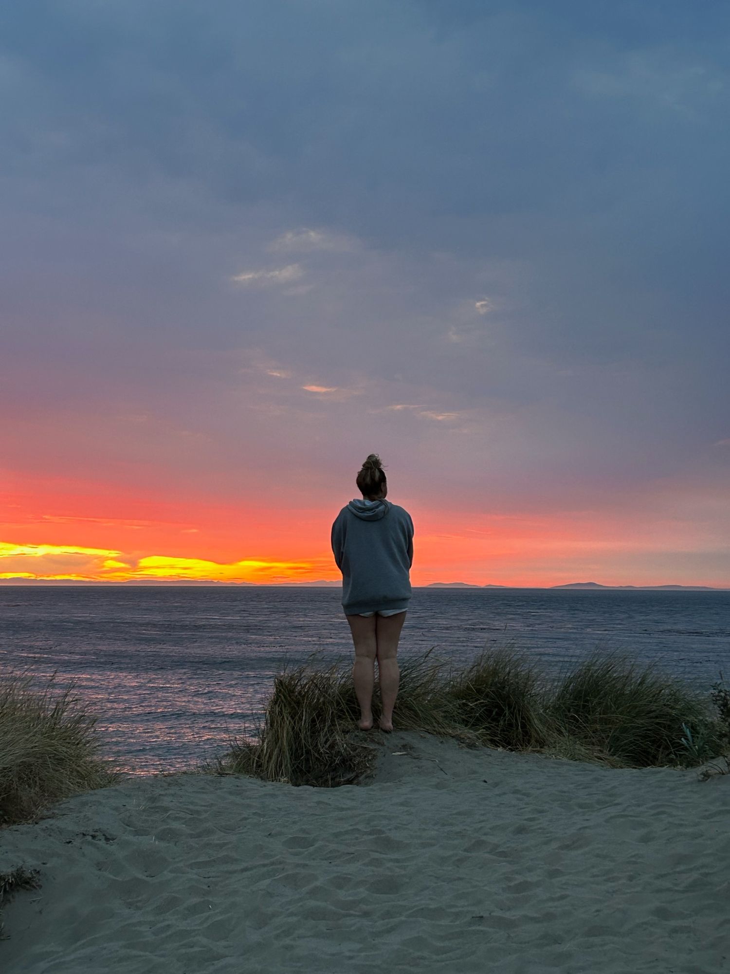Cora Emilie Media Silhouette stands on sandy dunes watching vibrant orange and pink sunset over ocean horizon.