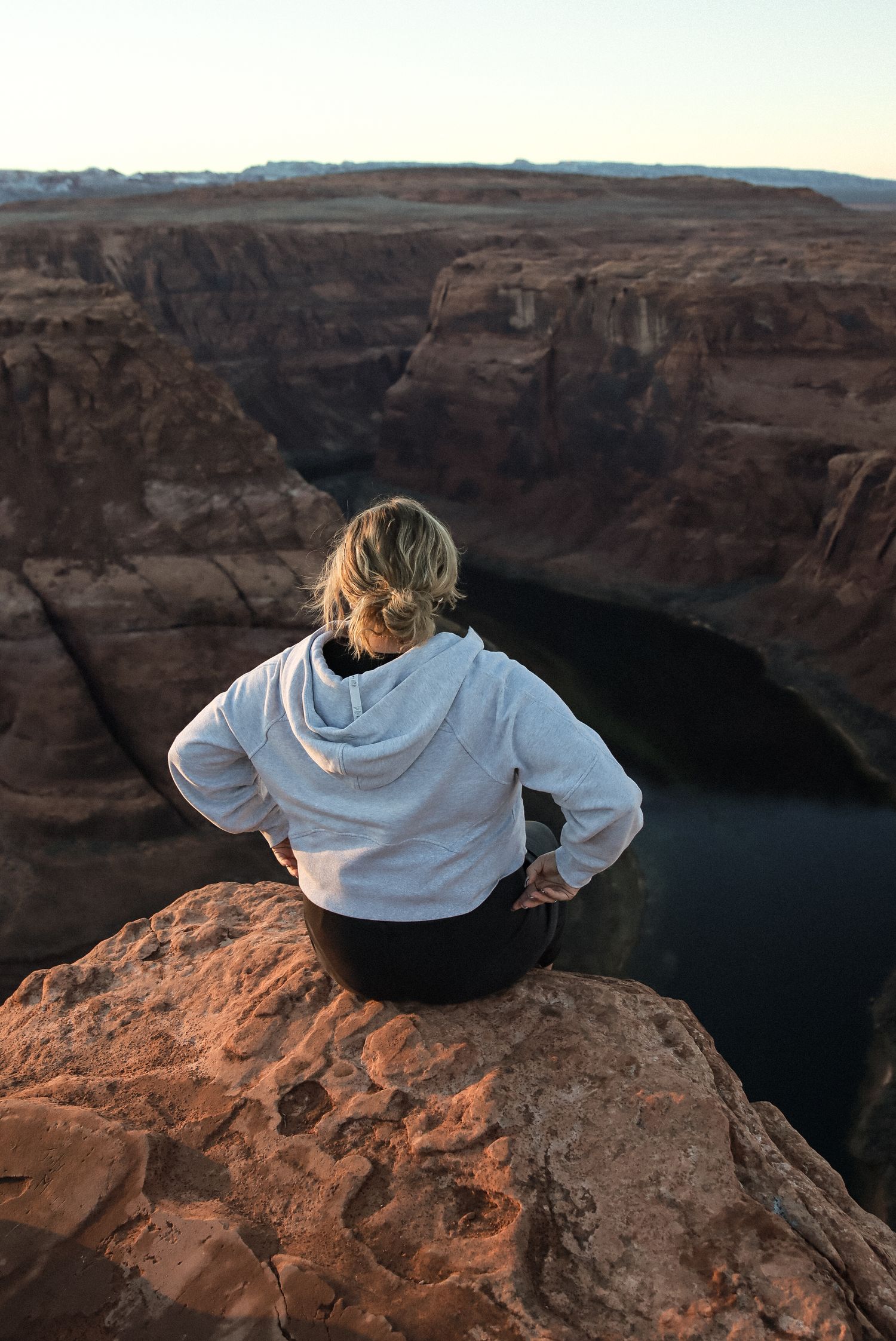 Cora Emilie Media Person in white hoodie sits on cliff edge overlooking dramatic canyon and dark river below.